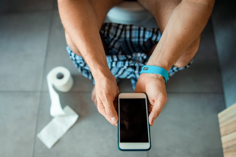 Imagen referencial en primer plano de un hombre sentado en el baño y sosteniendo un teléfono celular en sus manos.