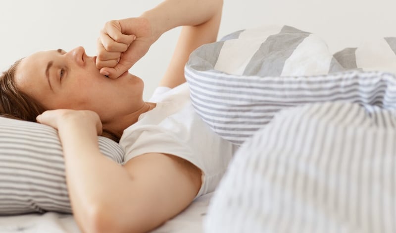 Mujer soñolienta con cabello oscuro con camiseta blanca casual acostada debajo de una manta y bostezando, tapándose la boca con el puño, simplemente despierta, disfrutando temprano en la mañana.